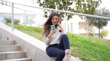 Genç bir kadın açık hava merdivenlerinde rahat bir şekilde oturuyor, akıllı telefonuyla canlı ve yemyeşil bir ortamda derinden meşgul, rahat ve keyifli bir açık hava yaşam tarzını gösteriyor.