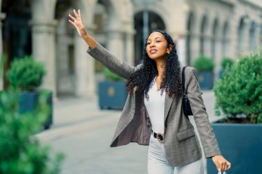Şık, kıvırcık saçlı genç bir kadın canlı bir şehir bölgesinde yürürken neşeli bir şekilde el sallıyor. Kusursuz moda anlayışını ve hayat dolu kişiliğini her adımında sergiliyor.