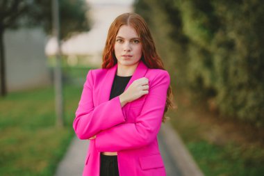 Yeşillik ve canlı renklerin sergilendiği bir açık hava ortamında, çarpıcı pembe bir ceket giymiş şık genç bir kadın, zarif ve modern bir kadınlık havası yayarak güvenli bir şekilde poz veriyor.