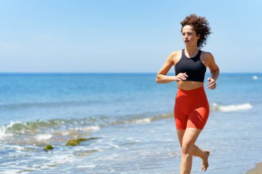 Dalgalı deniz suyunun yanındaki kumlu sahilde bulanık mavi gökyüzüne karşı gün ışığında koşarken spor kıyafetleri giyip gözlerini kaçıran, konsantre olmuş, kıvırcık saçlı genç bir kadın.