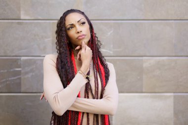 Bir kadın entrikalı dokulu bir duvara karşı düşünceli bir şekilde duruyor, kendine özgü örgülü saç stilini gözler önüne seriyor canlı kırmızı aksanlarla süslenmiş, kendine güveni, yaratıcılığı ve kişisel tarzını somutlaştırıyor.