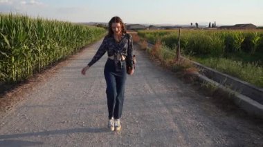 Bir kadın bereketli mısır tarlalarında dolambaçlı bir yolda yürüyor, çarpıcı bir günbatımının ılık güzelliğine dalmış, ruhu büyüleyen huzurlu ve huzurlu kırsal bir manzara yaratıyor.