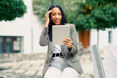 Genç bir kadın dışarıda keyifli bir anın tadını çıkarıyor. Tablet cihazıyla uğraşırken zarif bir şekilde bankta oturuyor. Parlak bir gülümseme sergiliyor.