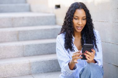 Canlı bir şehir ortamında, kıvırcık saçlı genç bir kadın açık hava basamaklarında oturuyor, akıllı telefonuyla tamamen meşgul, yoğun yaşamlarında modern yaşam tarzını sembolize ediyor