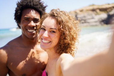 Kıvırcık saçlı kız arkadaşının yanında duran, yaz tatilinde kayalık kayalıklara ve okyanusa selfie çeken mutlu siyah adam.