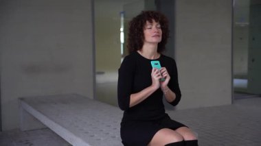 Kıvırcık saçlı kendine güvenen genç bir kadın modern bir akıllı telefon sergiliyor. Teknoloji ve moda yaşam tarzının karışımını vurgulayan canlı bir ortamla çevrili. Bu da onu ilgi odağı yapıyor.