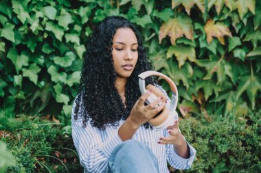 Genç bir kadın zarif kulaklıklarını huzur içinde inceliyor ve eğlenirken, aynı zamanda dingin ve gür yeşilliklerle çevrili, ki bu da dinginlik ve modern tarzı güzel bir şekilde somutlaştırıyor.