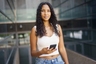 Gündelik kıyafet giymiş modaya uygun ve şık bir genç kadın şehir ortamında güven içinde duruyor. Akıllı telefonunu tutuyor ve bakışlarını kameraya çeviriyor, özgüven yayıyor.