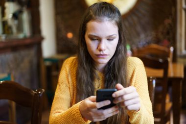 Sıcak ve rahat bir kafe ortamında, uzun dalgalı saçlı genç bir kadın rahat bir şekilde oturuyor, parlak sarı bir kazak giyiyor, tamamen akıllı telefonuna odaklanmış, dijital dünyasına odaklanmış.