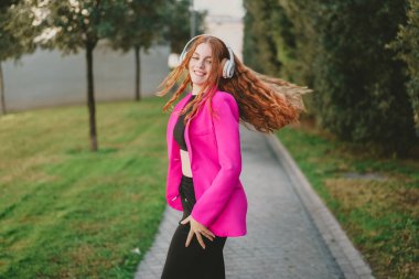 Şık pembe ceketli ve rahat kulaklıklı neşeli bir kadın, yemyeşil, mutluluk saçan, kendine güvenen ve kendine özgü tarzıyla çevrili büyüleyici bir yolda neşeyle dans ediyor.