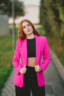 Kendine güvenen ve stil sahibi genç bir kadın zarif bir şekilde açık havada poz veriyor. Parlak pembe ceketini gösteriyor. Şık siyah bluzuyla zarif bir tezat oluşturuyor.