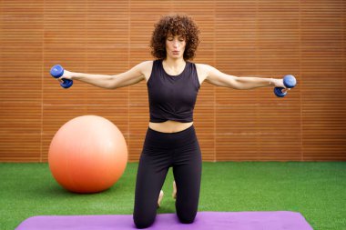Kendini adamış genç bir kadın yoga minderinin üzerinde dambıllarıyla omuz egzersizleri yapıyor, dengeli ve tatmin edici bir yaşam tarzı için zindeliğini, gücünü ve genel sağlığını arttırıyor.