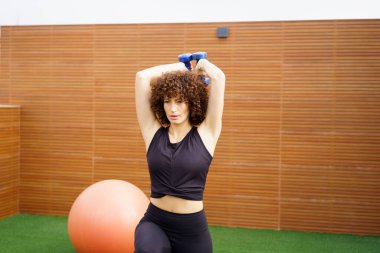 Güzel, kıvırcık saçlı bir kadın, hareketli bir egzersiz topu içeren modern ve şık bir spor salonunda, halterlerle güç egzersizleri yapıyor.
