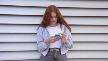 Kızıl saçlı genç bir kız, akıllı telefonuna kendini kaptırmış, güvenli bir şekilde şehir duvarının yanında duruyor, modern gençlik kültürünü ve günümüzde hızla gelişen dünyanın toplumsal etkilerini yansıtıyor.