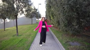 Şık, genç bir kadın, kendine güvenerek, şık, pembe bir ceket ve şık siyah bir pantolon giyerek, moda ve doğa dünyalarını güzel bir şekilde harmanlayarak yürüyor.