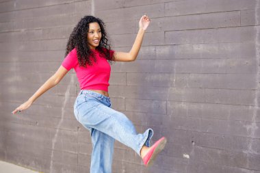 Neşeli pembe bluz ve modaya uygun bol kot giyen genç bir kadın neşeli bir gri duvara karşı neşe içinde poz veriyor, neşeli tarzını, bulaşıcı enerjisini ve renkli kişiliğini sergiliyor.