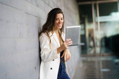 Genç bir kadın tabletini kullanırken parlak bir gülümseme sergiliyor, şık giyinmiş, kentsel bir ortamda duvara yaslanmış, rahat tavrını sergiliyor ve dijital etkileşimden zevk alıyor.