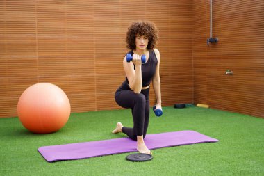 Formda ve güçlü bir kadın dambıllar kullanarak yoğun bir egzersiz yapar, spor aletleriyle dolu, canlı ve modern bir spor salonunda sağlık ve spor yapmaya olan bağlılığını gösterir.