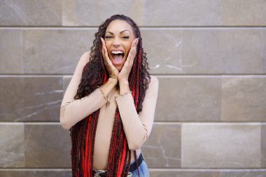 Neşeli ve coşkulu genç bir kadın coşkusunu ve mutluluğunu geniş, geniş bir gülümseme ve canlı, renkli örgülü saçlarıyla, şık ve modern şehir arkaplanına karşı sergiliyor.