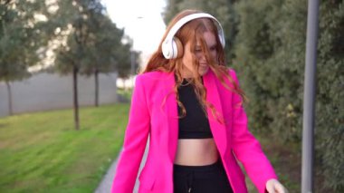 Parlak pembe bir ceket giyen ve kulaklık takan şık ve şık bir kadın, enerji ve neşe saçan canlı bir ortamda dışarıda vakit geçiriyor.
