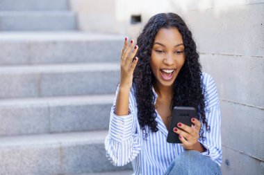 Güzel kıvırcık saçlı neşeli genç bir kadın akıllı telefonuyla bir dizi basamakta otururken heyecan verici bir şekilde etkileşime geçiyor. Mutluluğunu ve etrafındaki dijital dünyayla bağlantısını gösteriyor.