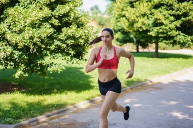 Yeşil bir parkta, atletik giyimli bir kadın koşar, güneşin altında spor ve sağlığa olan bağlılığını gösterir, egzersiz, sağlık ve açık hava aktivitesi gibi anahtar kelimeleri somutlaştırır.