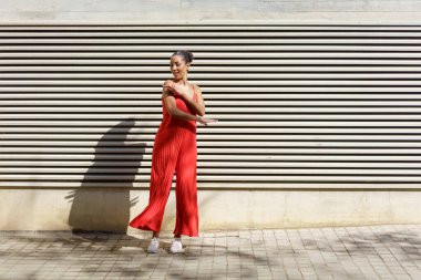 Kendine güvenen genç bir kadın, canlı kırmızı bir tulum giymiş, neşeli bir şekilde çizgili bir duvarın önünde poz veriyor, eğlenceli ruhunu ve moda ve tarz anlayışını sergiliyor.
