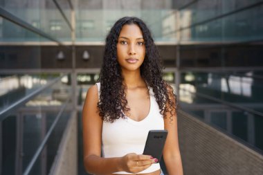 Modern bir ortamda, kendine güvenen kıvırcık saçlı genç bir kadın akıllı telefonuyla poz veriyor, yaratıcılık, moda ve çağdaş tarzdaki eğilimleri gösteriyor, yeni nesli somutlaştırıyor.