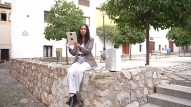 Resimli ve canlı bir plazada, taştan bir duvarda oturan bir kadın, tabletinin tadını çıkarırken, yemyeşil ağaçlarla çevrili ve manzarayı güzelleştiren büyüleyici, çağdaş bir mimariye sahip.