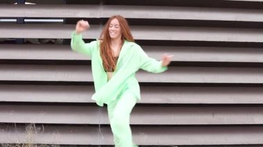 Canlı yeşil takım elbiseli bir kadın modern mimari bir arka plana karşı enerjik bir şekilde dans ediyor, neşe saçıyor ve kendine güvenen şık yeteneğini ve renkli ifadesini sergiliyor.