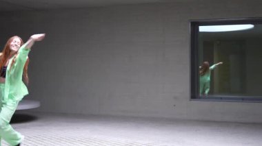 Kızıl saçlı bir kadın, parlak yeşil bir kıyafetle, minimalist bir kapalı alanda enerjik bir şekilde dans ediyor. Yansıması bir pencerede görülüyor. Bu dinamik ve anlamlı performans enerji yayar.