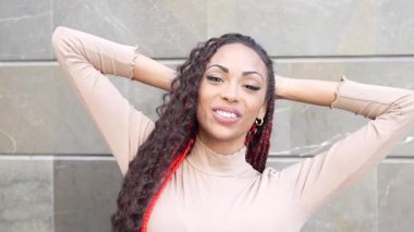 Uzun örgülü saçları olan neşeli, hayat dolu bir kadın parlak bir gülümseme sergiliyor ve modern mimari bir duvarın arka planında kendine güvenle poz veriyor, büyüleyici ve pozitif enerji yayıyor.