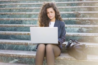 Kıvırcık saçlı genç bir profesyonel açık hava basamaklarında oturuyor ve bilgisayarına odaklanmış durumda. Zarif giyinmiş, yanında şık bir çanta, verimliliği ve modern çalışma kültürünü somutlaştırıyor..
