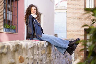 Canlı ve büyüleyici bir şehir arka planında, doğal kıvırcık saçlı, modaya uygun deri ceket giymiş, mutlu mesut boş zamanlarının tadını çıkaran, taş bir duvarda rahat bir şekilde oturan,