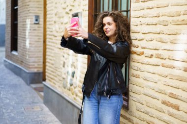 Güzel kıvırcık saçlı, şık ve şık bir genç kadın canlı ve çekici bir selfie için heyecanlı bir çevrede dokulu bir şehir duvarına karşı poz veriyor ve bir anlık neşe yakalıyor.