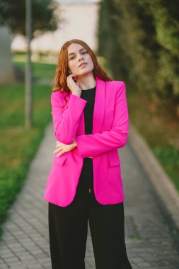 Kendine güvenen genç bir kadın kendine güvenen parlak pembe bir ceketin içinde duruyor. Parlak, siyah bir pantolonu var. Canlı bir şehir arkaplanına karşı mükemmel bir şekilde çağdaş moda sergiliyor.