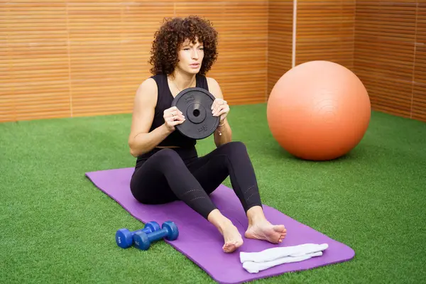 Kendini adamış ve odaklanmış bir kadın, vücut geliştirme eğitimine katılır, ağırlık plakasını kaldırır, spor salonundaki çeşitli spor aletlerinin arasında, spor yapmaya olan bağlılığını vurgular.