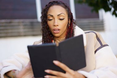 Odaklanmış bir kadın, çağdaş yaşam tarzını yansıtan şık ve şık bir kıyafet içinde modern bir okuma deneyimini sergileyerek açık havada bir kitap okumaya dalmıştır.