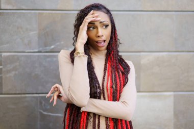 Şık bir kadın canlı kırmızı ve siyah rastalarını gururla sergilerken modern bir duvar zeminine karşı düşünceli bir poz vererek şık görünüşünü tamamlıyor ve çerçeveliyor.