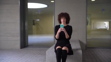 Kıvırcık saçlı genç bir kadın, çağdaş tasarım ve kentsel yaşam tarzının bir karışımını barındıran gösterişli ve modern bir mekanda otururken akıllı telefonuna kendini kaptırmış bir şekilde taş bir bankta oturuyor.