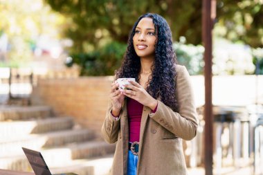 Genç bir kadın dışarıda, güzel bir ortamda elinde bir kahve fincanı, neşe ve pozitiflik saçarak duruyor. Huzurla çevrelenmiş bir anlık tefekküre dalmış.
