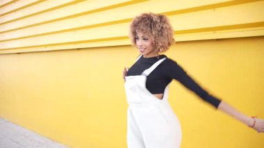 Canlı ve coşkulu bir kadın saf özgürlüğü kucaklıyor, parlak sarı bir duvarın önünde neşeyle dans ediyor, parlak yaratıcılığını ve dizginlenemez mutluluğunu gösteriyor.