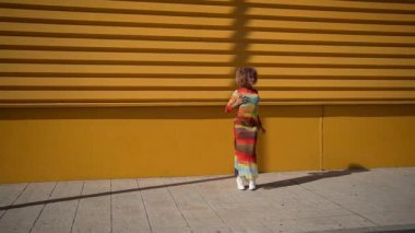 Şık bir birey kendine güvenerek canlı sarı bir duvara karşı poz veriyor, renkli bir kıyafet sergiliyor, dikkat çeken ve pozitif ışık saçan neşeli bir tutum sergiliyor.