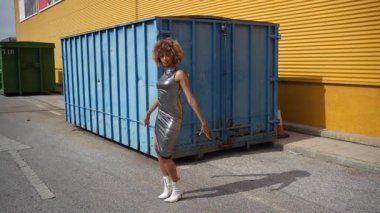 Şık bir birey, canlı nakliye konteynırlarının zeminine çarpıcı bir şekilde yerleştirilmiş parlak metalik bir elbisenin içinde, modern şehir modasının özünü güzel bir şekilde gözler önüne seriyor.