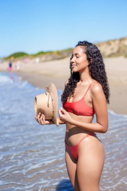Canlı kırmızı bir bikini giymiş genç bir kadın zarif bir şekilde elinde şık bir hasır şapka tutarak güneşli plajın keyifli atmosferinin keyfini çıkarıyor.
