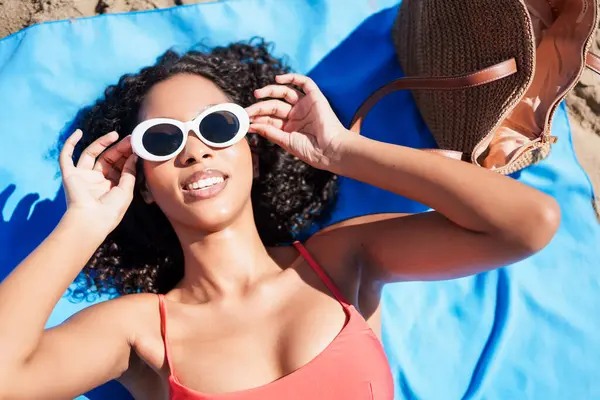 Genç bir kadın renkli bir plaj havlusunda rahatça dinleniyor, sıcak güneşin altında güneşleniyor, son moda güneş gözlüklerini gösteriyor ve mutluluk ve güzellik saçan kaygısız, neşeli bir ruhun tadını çıkarıyor.