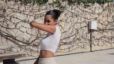 Temiz çörek saç stiline sahip neşeli ve enerjik bir kadın açık havada güneşli bir günün keyfini çıkarıyor. Zarif beyaz bir bluz giyer ve zarif bir şekilde eşsiz desenli bir duvara poz verir.