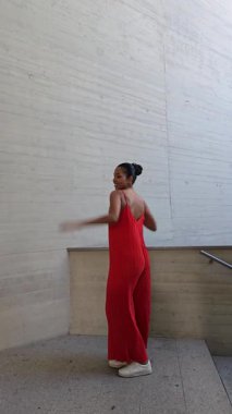 Dalgalı kırmızı elbiseli neşeli genç bir kadın modern moda ve yaratıcılığı harmanlayan şehir ortamında neşeli bir poz veriyor. Neşeli tavırları mutluluk saçıyor ve güven veriyor.