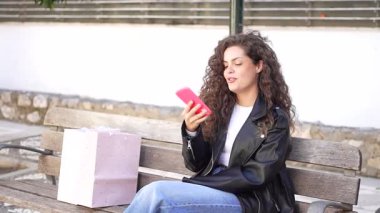 Kıvırcık saçlı genç bir kadın düşünceli bir şekilde bir bankta oturuyor, yanında şık bir alışveriş çantasıyla telefonunu tutuyor, güzel bir günde huzurlu bir rahatlama anının tadını çıkarıyor.