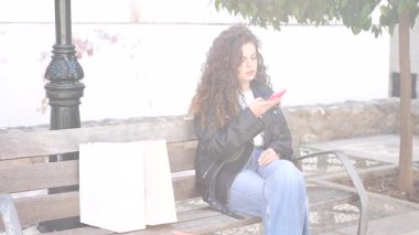 Güzel kıvırcık saçlı, genç bir kadın, bir park bankında rahatça oturuyor, tamamen akıllı telefonuna dalmış, çevresi yeşilliklerle doluyken.